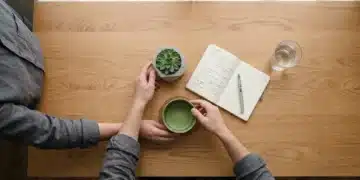 An aerial view of a person setting down a green matcha bowl next to an open journal and a succulent on a minimalist wooden desk, representing a structured morning routine for focus and clarity.